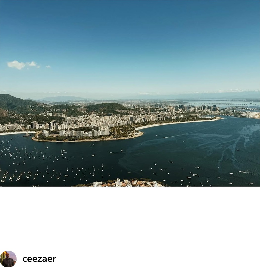 Event Coverage - Aerial view of Guanabara Bay with hundreds of sailboats, coastal cityscape, forested mountains, and a long bridge stretching across the water under clear blue skies