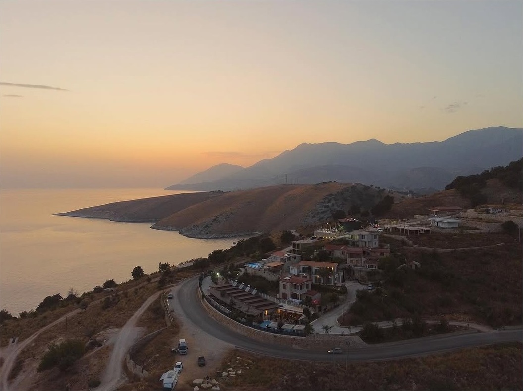 Sunset Sessions - Aerial drone view of a coastal Mediterranean village at golden hour with winding roads, hillside buildings, and calm sea reflecting the warm sunset glow